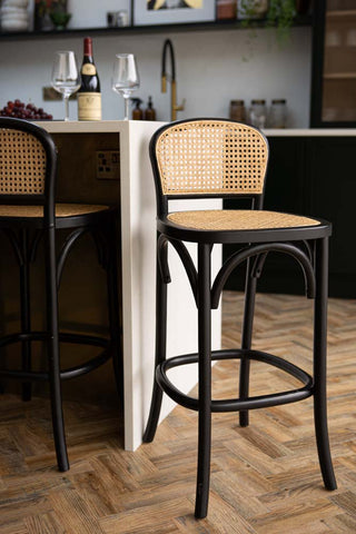 The Chez Pitou Black Wood & Woven Cane Bar Stool displayed in a kitchen in front of a breakfast bar.