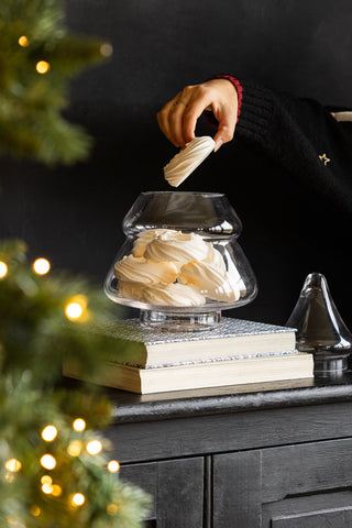 The Christmas Tree Glass Storage Jar on a sideboard on some books, with the lid off and a model taking a meringue from inside.