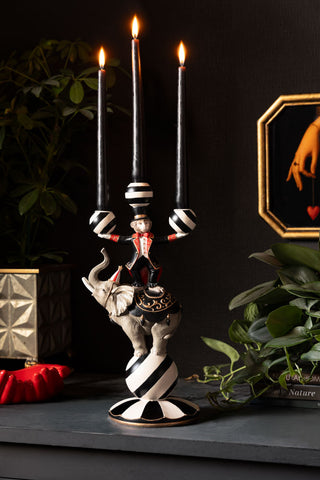 The Circus Animal Acrobat Candelabra styled with three lit black candles, displayed on a dark sideboard with a plant and various accessories.