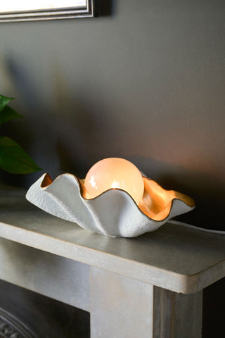 The Clam Table Lamp illuminated and displayed on a fireplace shelf.