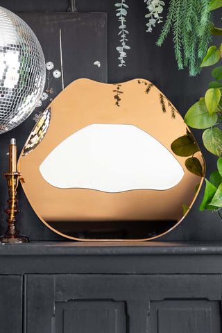The Copper Pink Lips Decorative Mirror displayed on a black sideboard, styled with plants, an amber candlestick holder and a silver disco ball.