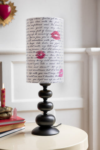 The Curve Black Table Lamp With Love Letter Shade shown on a white side table against heart and lips home accessories and a white panelled wall. 