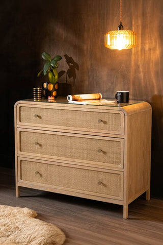 The Curved Wood & Rattan Chest Of Drawers styled with a plant and various accessories on, in front of a dark wall.