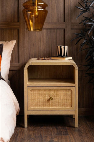The Curved Wood & Rattan Bedside Table styled next to a bed, with some accessories on/around it.