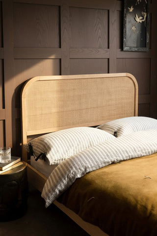 The Curved Wood & Rattan Bed styled with bedding on, with a bedside table and framed artwork also in shot.