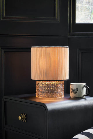 Lifestyle image of the Amber Parisian Table Lamp displayed illuminated on a side table with a mug. 