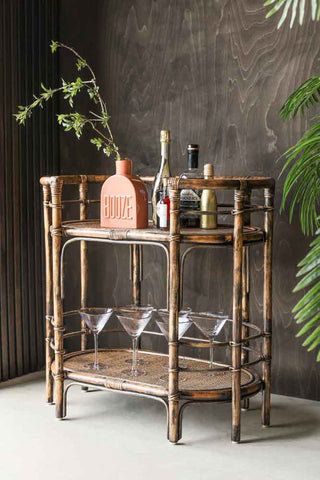 Lifestyle image of the Dark Brown Rattan Console/Drinks Table styled with glasses and bar accessories against a dark wood wall with plants.