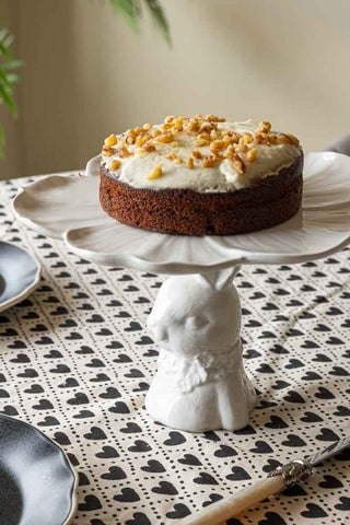 Lifestyle image of the Decorative Rabbit Cake Plate styled on a heart patterned tablecloth and a carrot cake on top. 