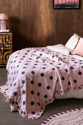 The Dot Noir Ruffled  Bedspread shown against a textured wood wall with rattan and pink accessories. 