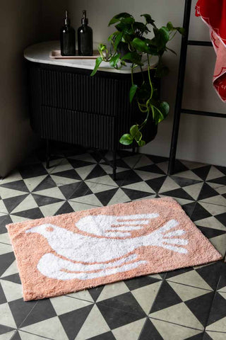 The Dove Tufted Cotton Bath Mat styled on a monochrome patterned floor in front of a storage cupboard, with soap dispensers, plant and a towel rack with a towel on.
