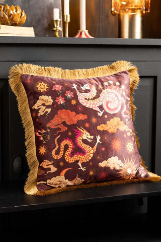 The Dreamland Dragon Fringed Velvet Cushion on a bench in front of a black sideboard.