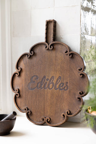 The Edibles Scalloped Wooden Serving Board shown against white zellige wall tiles in a kitchen. 