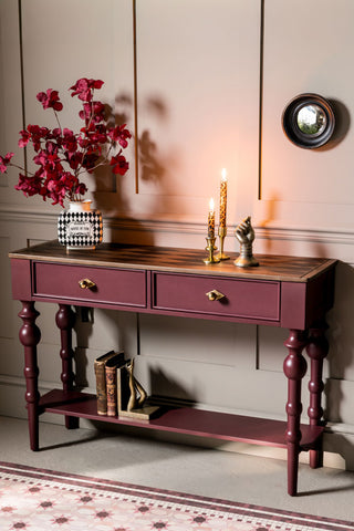 The Emporium Brick Red Console Table shown in a hallway with panelled walls, styled with books, leopard candles and a vase with faux flowers. 
