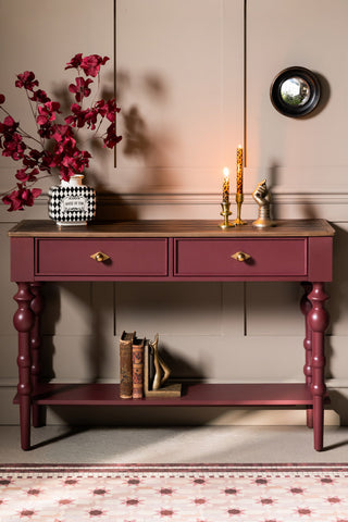 The Emporium Brick Red Console Table, shown straight on against beige panelled walls with a vase and leopard candles on the top and vintage books on the bottom shelf. 