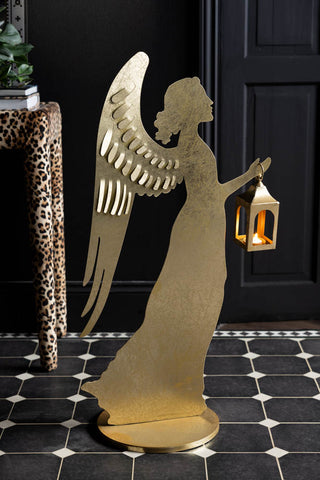 The Extra Large Gold Angel Decorative Lantern with a lit candle inside, displayed on a geometric floor with a leopard print table in the background.