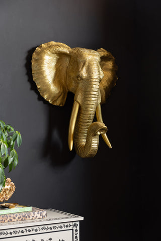 The Extra Large Gold Elephant Head Wall Art shown against a dark black wall with a plant and folk chest of drawers in the background.