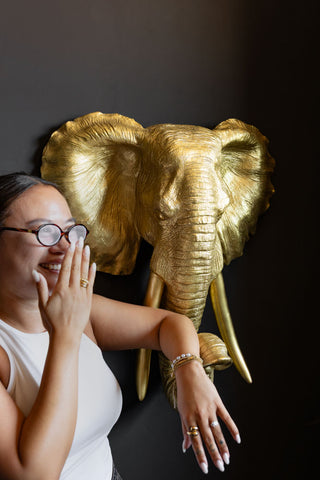 A scale photo of the The Extra Large Gold Elephant Head Wall Art, with a woman stood next to the elephant head to give an idea on scale. 