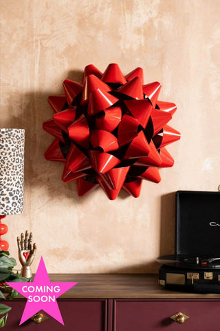 The Large Red Gift Bow Christmas Decoration styled on the wall above a sideboard with various accessories on. Image is shown with a pink icon that reads 'coming soon'.