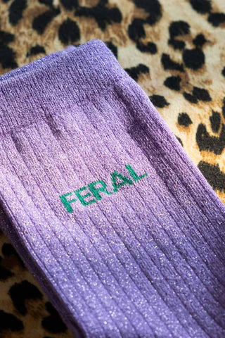The Feral Cotton Glitter Socks shown against a leopard background. Socks are purple with green writing that says 'feral'. 