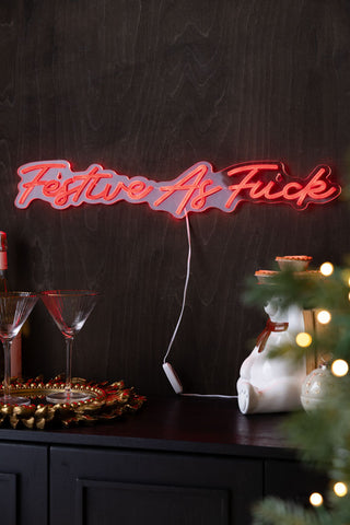 Neon sign with 'Festive As Fuck' on a dark background with Christmas decor.