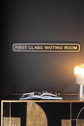 Lifestyle image of First Class Waiting Room Sign, displayed on a black wall above a gold table with an illuminated lamp and book. 