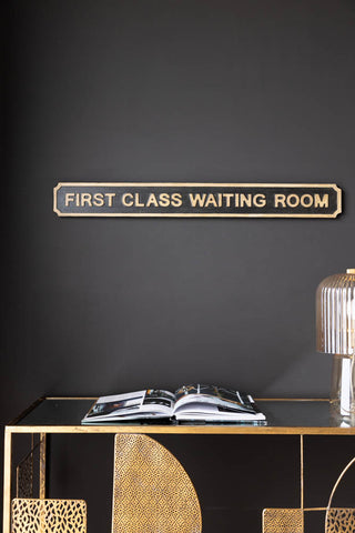 Lifestyle image of First Class Waiting Room Sign, displayed on a black wall above a gold table with  a lamp and book. 