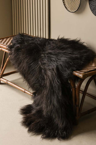 Lifestyle image of the Genuine Icelandic Long Wool Sheepskin - Natural Black, styled on a wicker bench in the corner of a neutral room. 