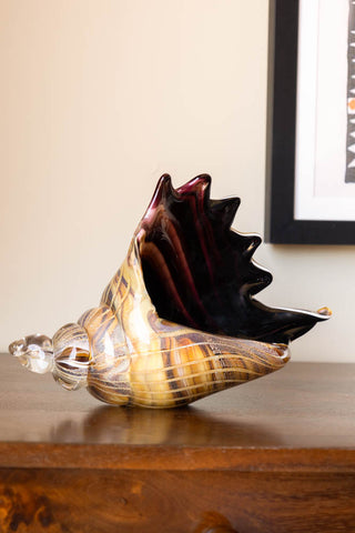 The Glass Conch Shell Paperweight Ornament on a wooden surface, with an art print on the wall in the background.