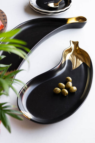 The Gold Aubergine Serving Bowl displayed on a white table with the Gold Banana Serving Bowl, styled with plates, cutlery and food.