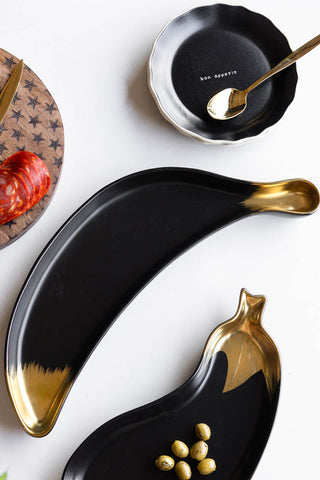 The Gold Banana Serving Bowl displayed on a white table with other kitchenware.