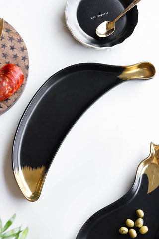 The Gold Banana Serving Bowl displayed on a white table with other kitchen accessories, styled with some olives and meat.