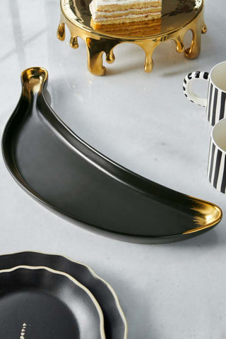 The Gold Banana Serving Bowl displayed on a marble worktop with mugs, plates and a cake stand.