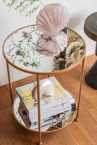 Lifestyle image of the Gold Beautiful Birds Mirror Side Table styled with decorative accessories and magazines. 