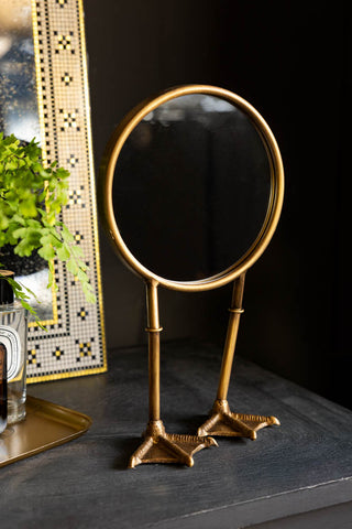 Gold Bird Legs Tabletop Mirror shwon against black walls with gold accessories. 
