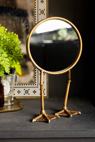 The Gold Bird Legs Tabletop Mirror shown on a black table with a gold tray and foxed mirror in the background.