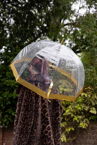 The Clear & Gold Domed Umbrella shown with a model wearing a leopard coat, outside in the rain.