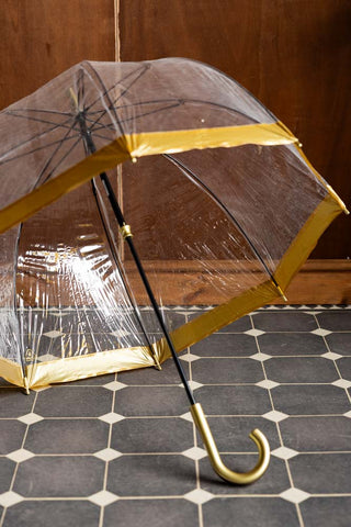 The Clear & Gold Domed Umbrella shown open inside against a wood wall and tiled floor mat. 