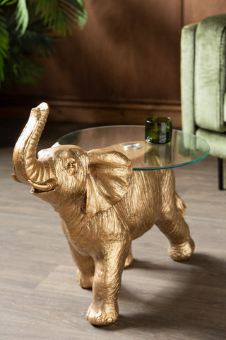 The Gold Elephant Side Table With Glass Top styled with a green glass on, with a sofa and plant in the background.