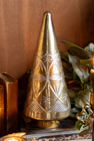 The Gold Etched Christmas Tree Display Ornament displayed with some books, greenery and lit candle in front of a wooden background.