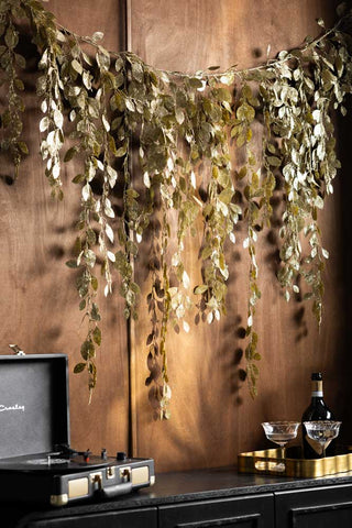 The Gold Glitter Eucalyptus Leaf Christmas Garland styled on a dark wall above some accessories on a black sideboard.