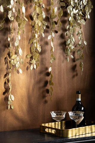 The Gold Glitter Eucalyptus Leaf Christmas Garland hanging in front of a dark wall above a tray, glasses and a bottle.