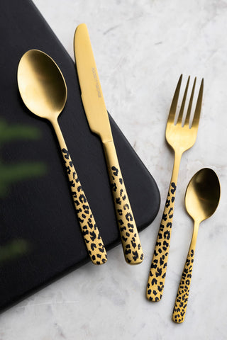The Gold Leopard 16 Piece Cutlery Set, including a spoon, knife, fork and teaspoon shown styled on a black wooden bread board on a marble counter. 