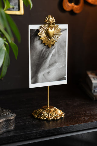 Gold Milagro Heart Photo Stand shown on a black sideboard with a black and white postcard on display. 