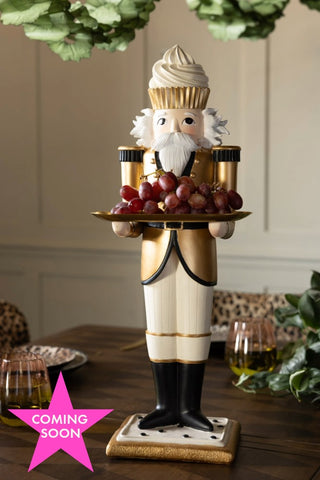 The Gold Nutcracker Butler Christmas Decoration holding some grapes on a table, with a pink star with the words 'coming soon' on in the bottom left corner.