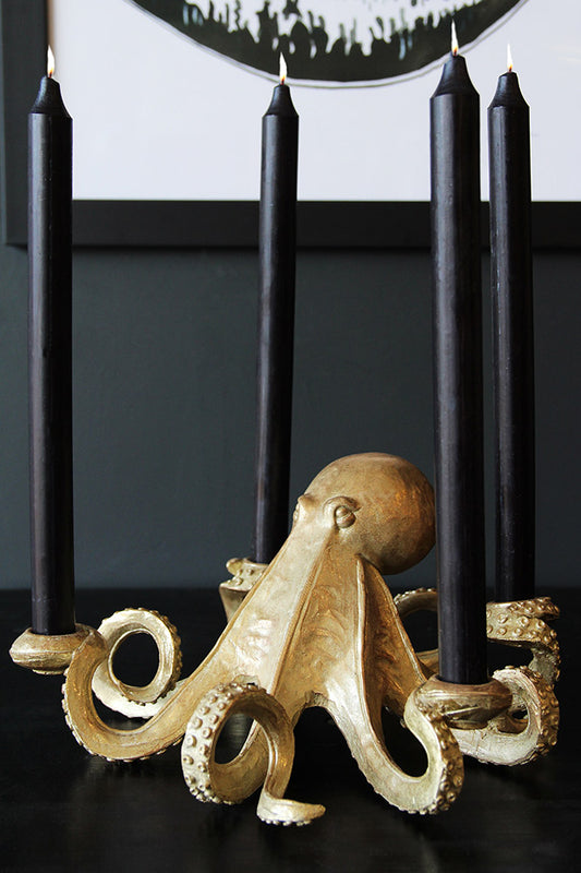 The Gold Octopus Candlestick Holder styled with tall black lit candles inside, displayed on a dark surface with an art print in the background.