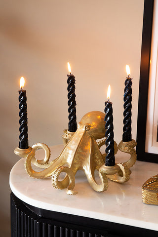 The Gold Octopus Candlestick Holder styled with lit black candles inside, displayed on a marble surface with an art print and gold ornament.
