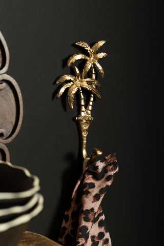 The Gold Palm Trees Wall Hook styled on the wall with a leopard print tea towel hanging on it, displayed next to a shelf with some accessories on.