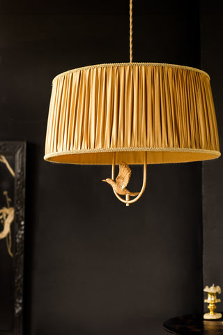 The Gold Ruched Hanging Bird Pendant Light shown with the light off, against a black wall with gold accessories in shot. 