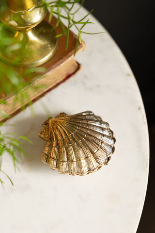 The Gold Shell Trinket Dish with the lid closed, displayed on a white table with a book, candle holder and a plant.