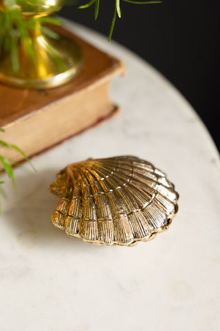 Close-up of the Gold Shell Trinket Dish with the lid closed, displayed on a white surface with a book, candle holder and book.